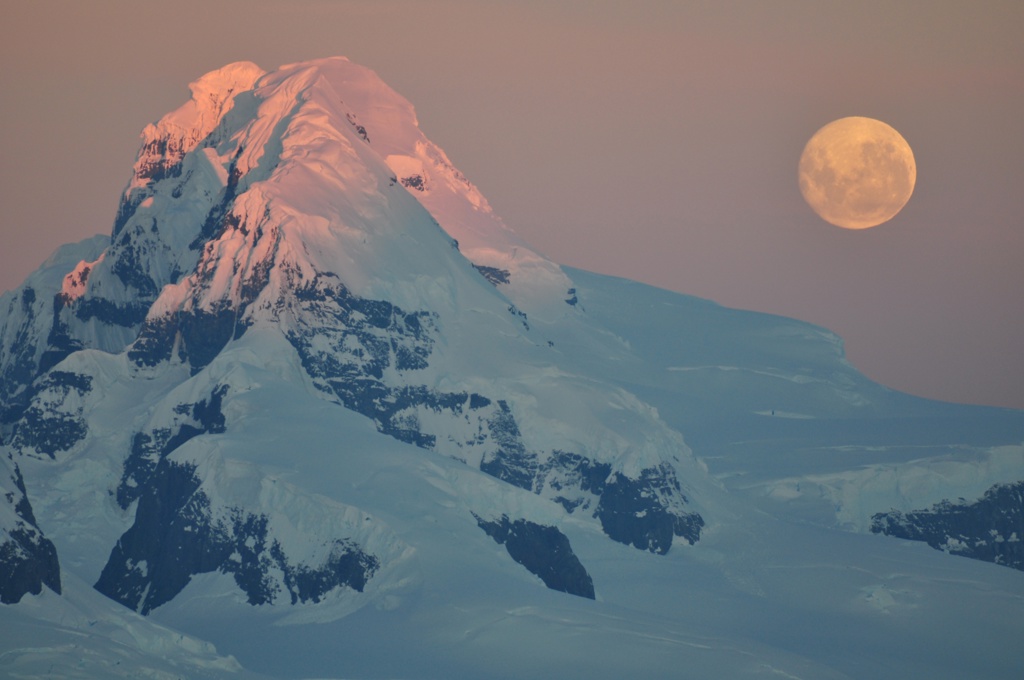 Besneeuwde bergtop in Antarctica bij zonsondergang met volle maan.