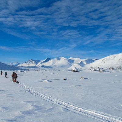 Langlaufers trekken met rugzakken over een besneeuwde vlakte richting bergtoppen onder een blauwe lucht.