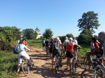 Fietstocht met groep reizigers over een zandpad in Myanmar, met een tempeltoren in de verte.