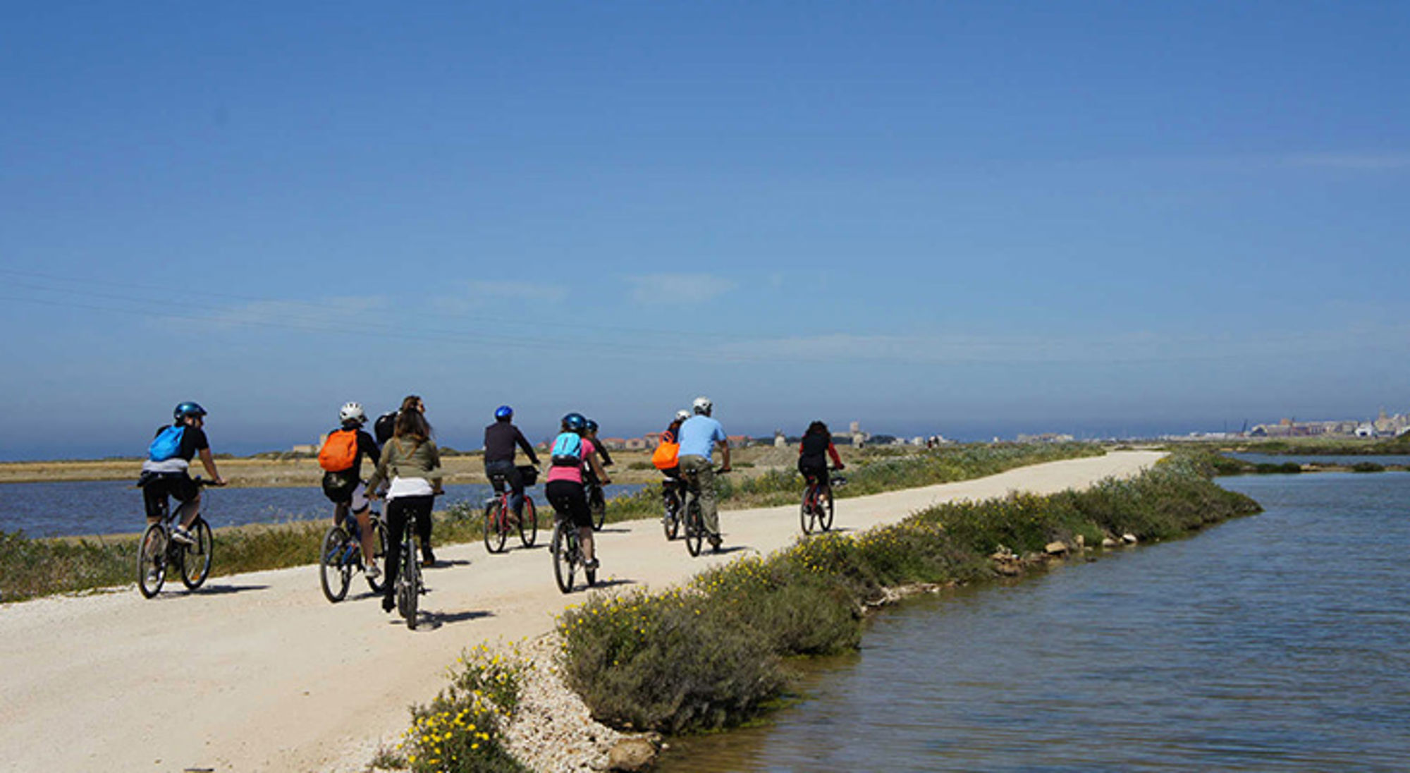 Groep fietsers op een kustpad langs water en duinen onder een heldere blauwe lucht.