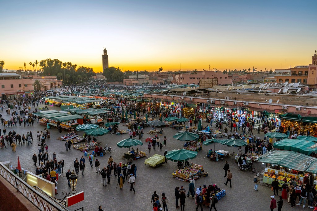 Drukke Djemaa el Fna-markt in Marrakech bij zonsondergang, met kraampjes en de Koutoubia-moskee op de achtergrond.