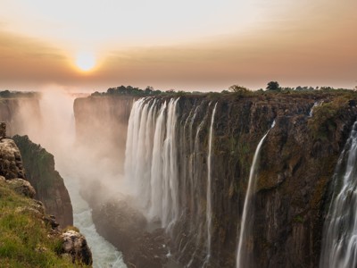 Victoria Falls bij zonsondergang, met nevel boven de kloof en watervallen langs de rotswand.