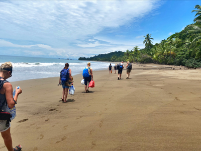 Wandelaars met rugzakken op een tropisch strand aan de Costa Ricaanse kust, met palmbomen en oceaan op de achtergrond.