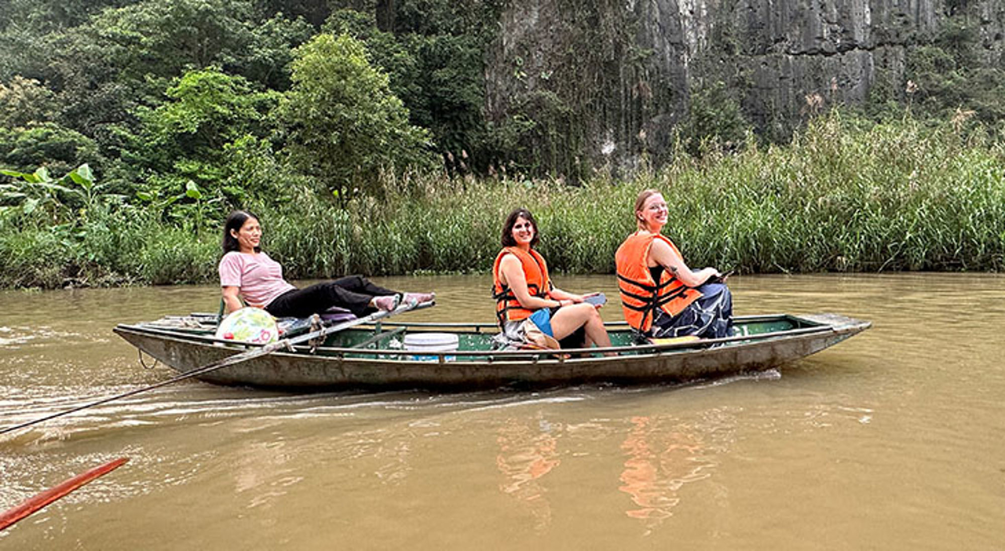 Boottocht Vietnam Drie reizigers met reddingsvesten in een boot op een rivier langs groene oeverbegroeiing.