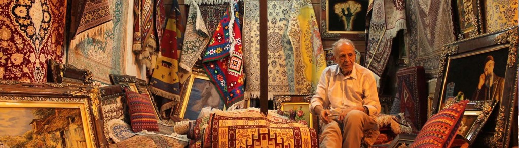 Tapijtenverkoper tussen kleurrijke Perzische tapijten en kussens op een bazaar in Iran.
