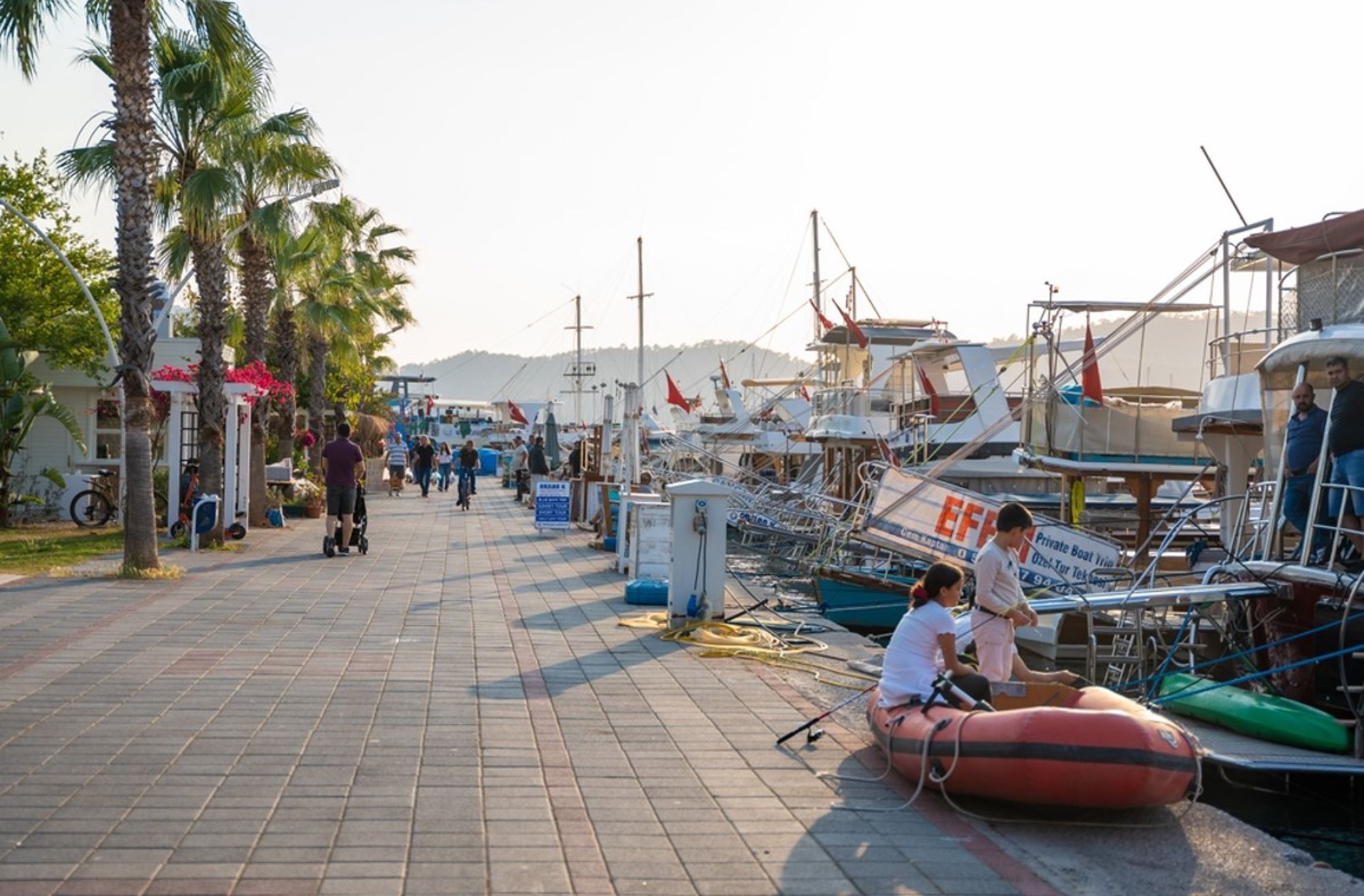 Promenade langs de jachthaven in Turkije met palmbomen, aangemeerde boten en wandelaars.