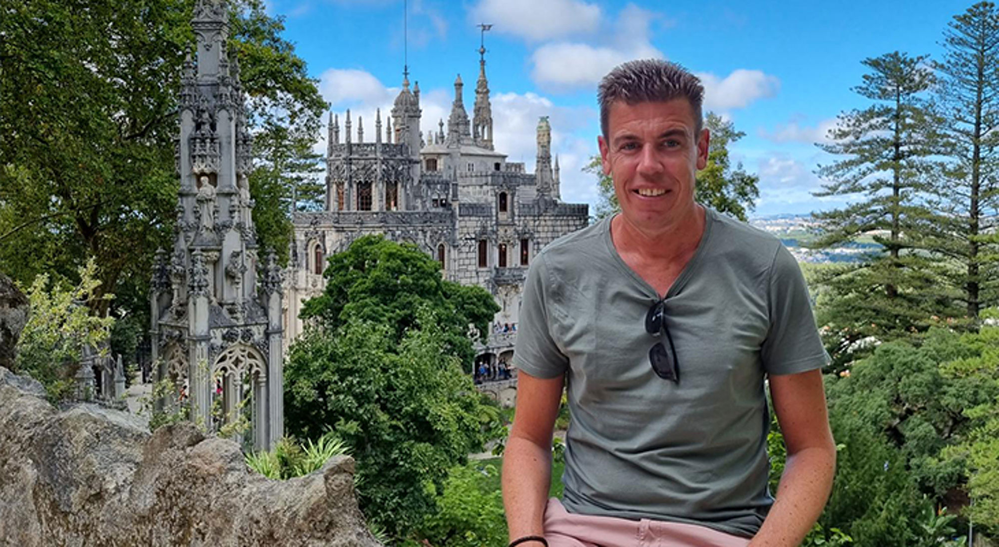 Jesse Man poseert bij het Pena Palace in Sintra, Portugal, met het kasteel en groene tuinen op de achtergrond.