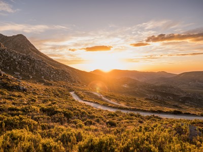 Zonsondergang boven een slingerende bergweg in Zuid-Afrika, met rotsachtige heuvels en struikgewas.