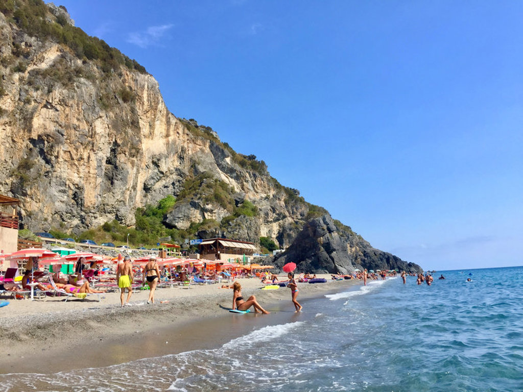 Druk strand met parasols aan de Amalfikust in Zuid-Italië, met hoge rotskliffen en helderblauwe zee.