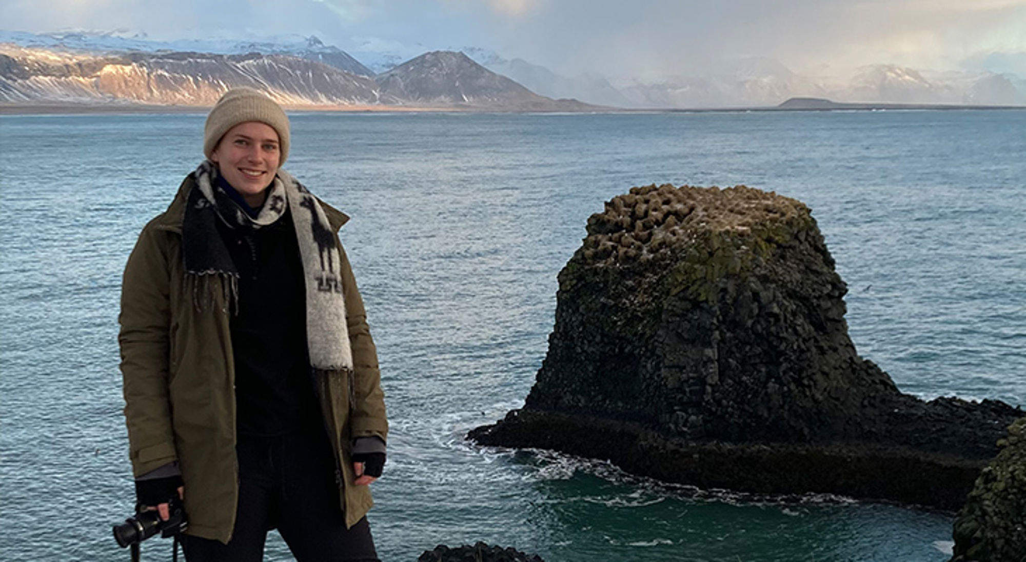 Carolien in IJsland Reiziger aan de kust met basaltrots en besneeuwde bergen op de achtergrond.