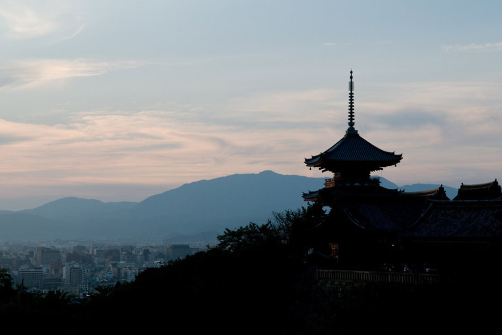 Silhouet van een Japanse tempel met pagode bij zonsondergang, met bergen en stad op de achtergrond.