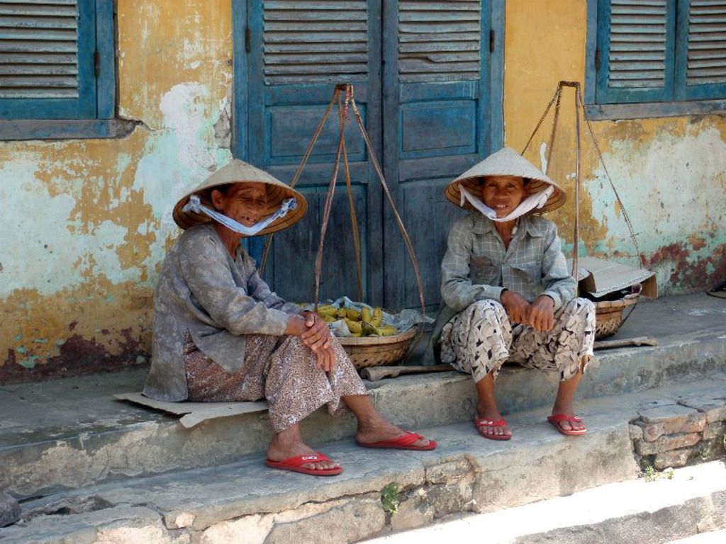 Twee Vietnamese vrouwen met rieten hoeden zitten bij een mand met fruit voor een blauw houten huis.