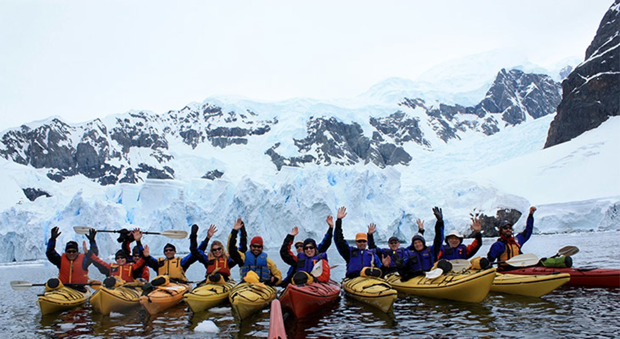 Groep kajakkers op het water met ijsbergen en besneeuwde bergen op de achtergrond.