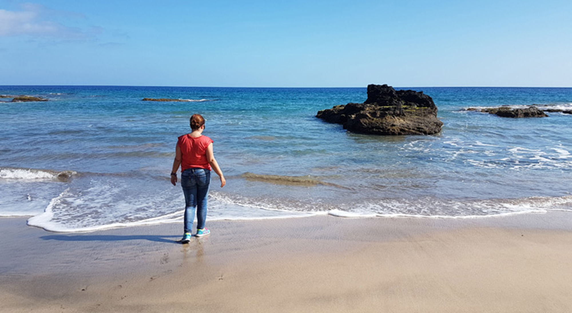 Vrouw wandelt langs de vloedlijn op een rustig zandstrand met rotsen in zee.