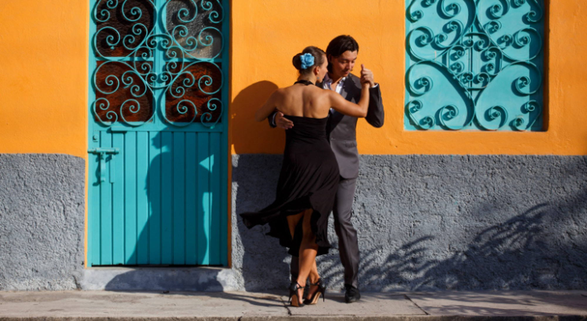 Dansend tango-paar op straat voor een feloranje muur met turquoise deur en sierlijke raamhekken.
