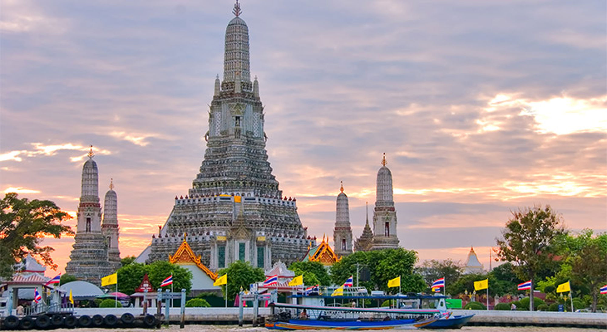 Wat Arun-tempel in Bangkok bij zonsondergang, met boten op de Chao Phraya-rivier en Thaise vlaggen.