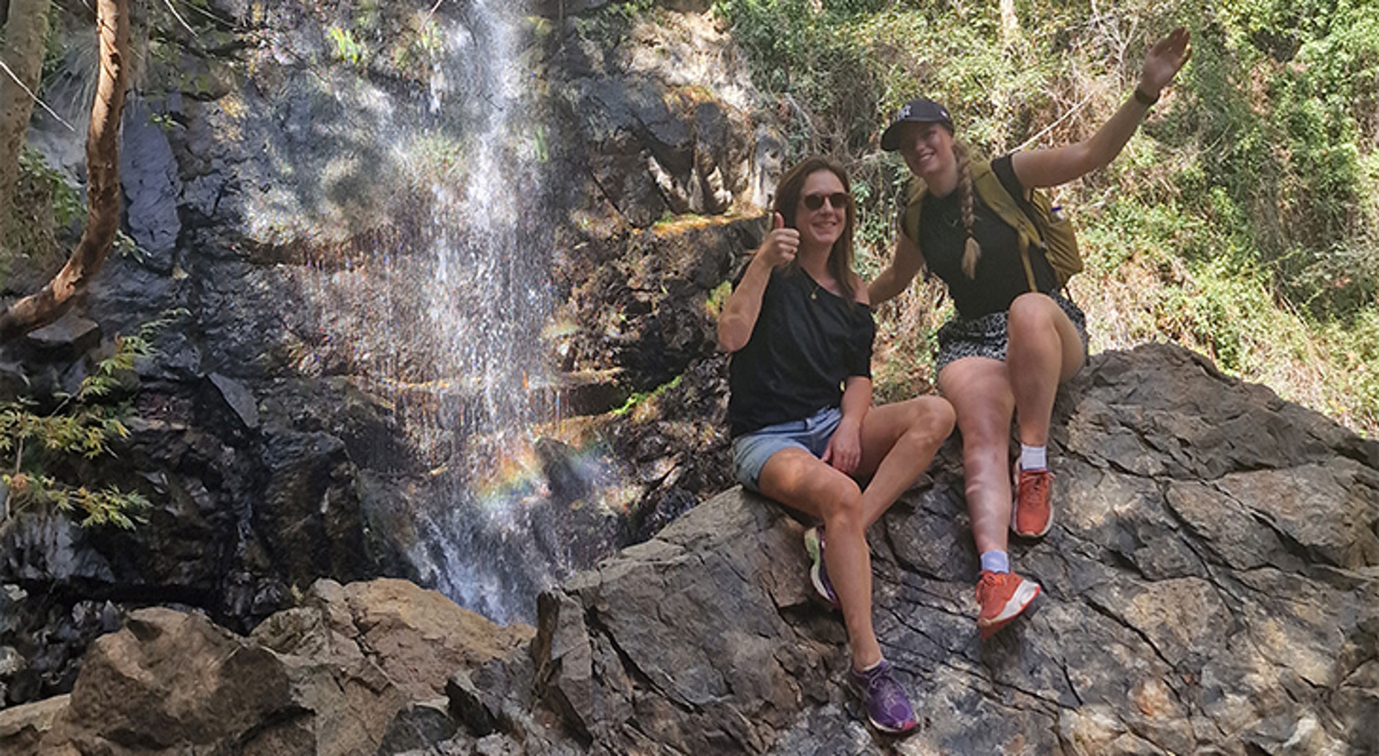 Twee wandelaars poseren op een rots bij een waterval in een bosrijke omgeving.