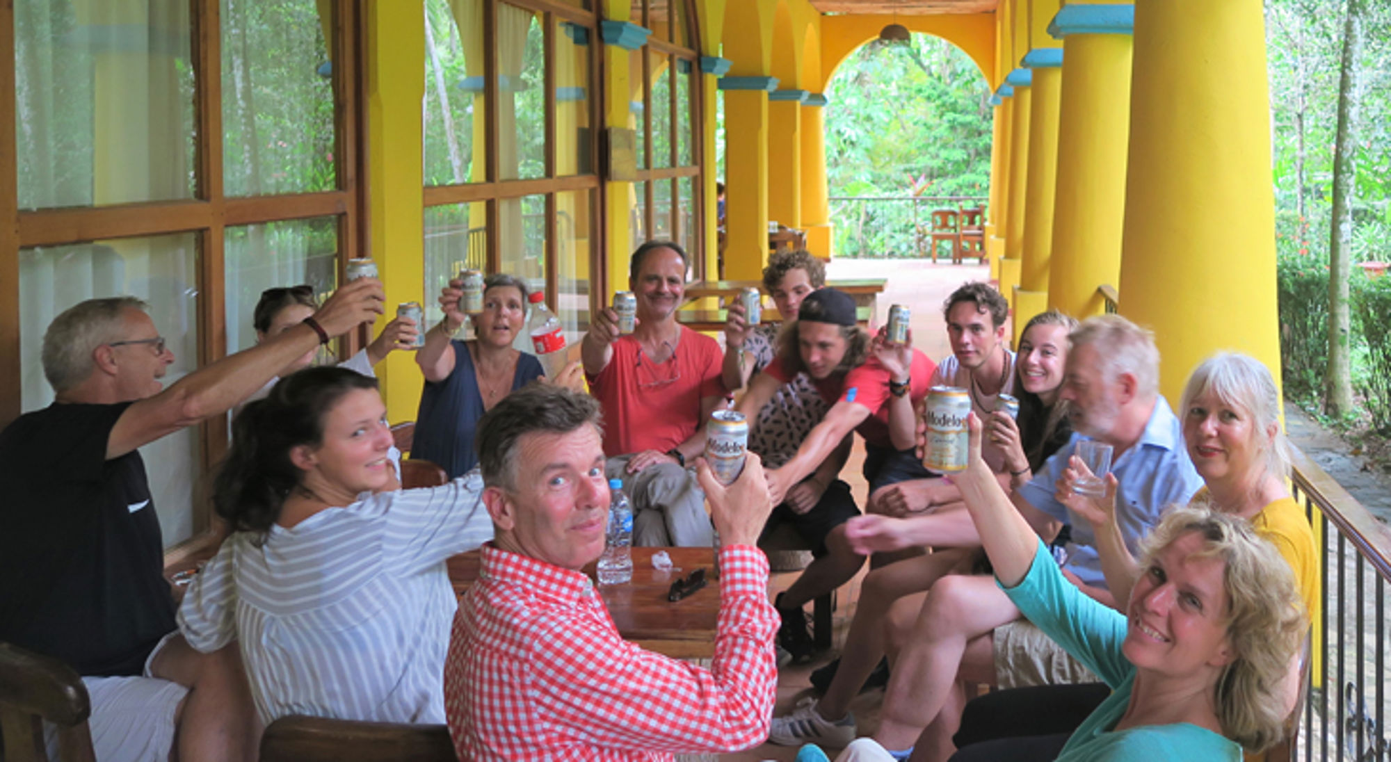 Groep reizigers proost met drankjes op een veranda met gele zuilen, omringd door groen.