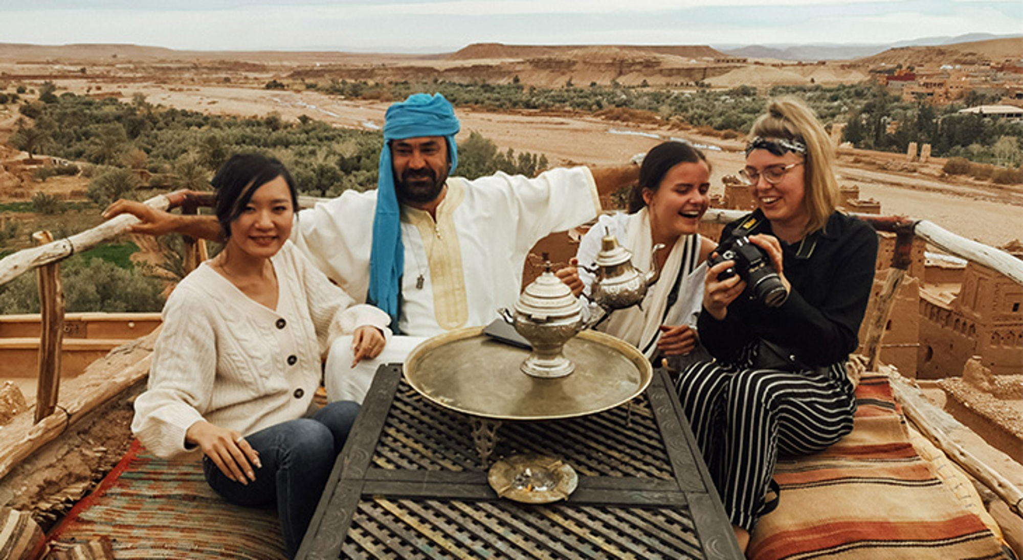 Foto met touristen Aït-Ben-Haddou Reizigers drinken muntthee op een dakterras met uitzicht op een woestijnvallei en lemen gebouwen.