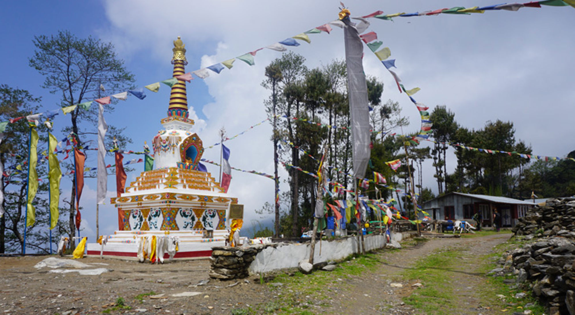 Boeddhistische stupa met kleurrijke gebedsvlaggen langs een bergpad bij een klein gebouw.