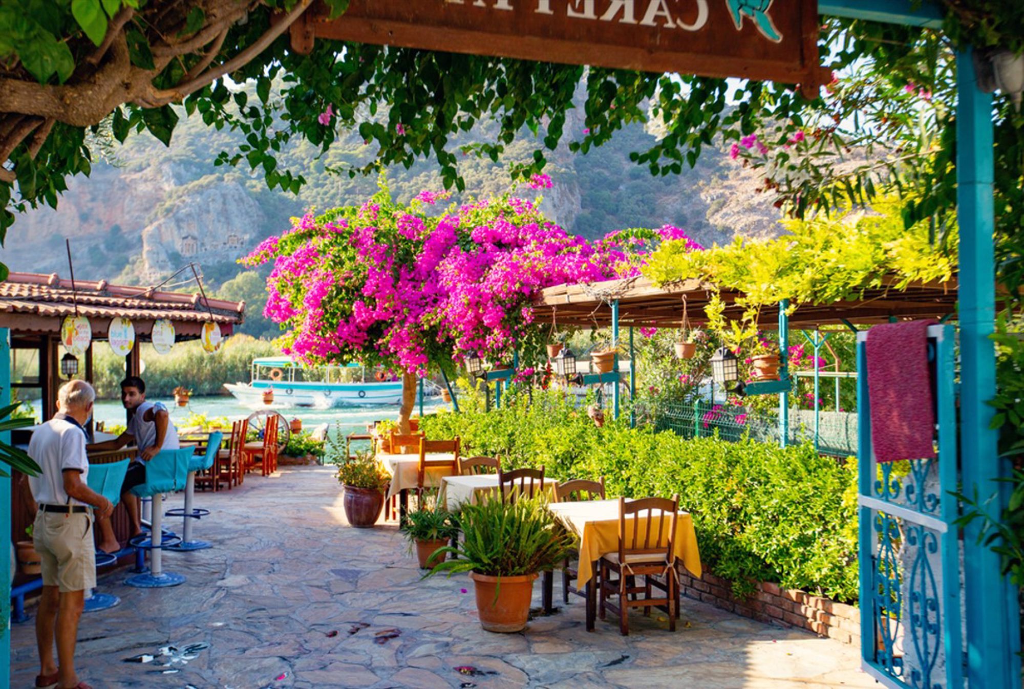 Terras aan het water in Turkije met kleurrijke bougainvillea, boten en bergen op de achtergrond.