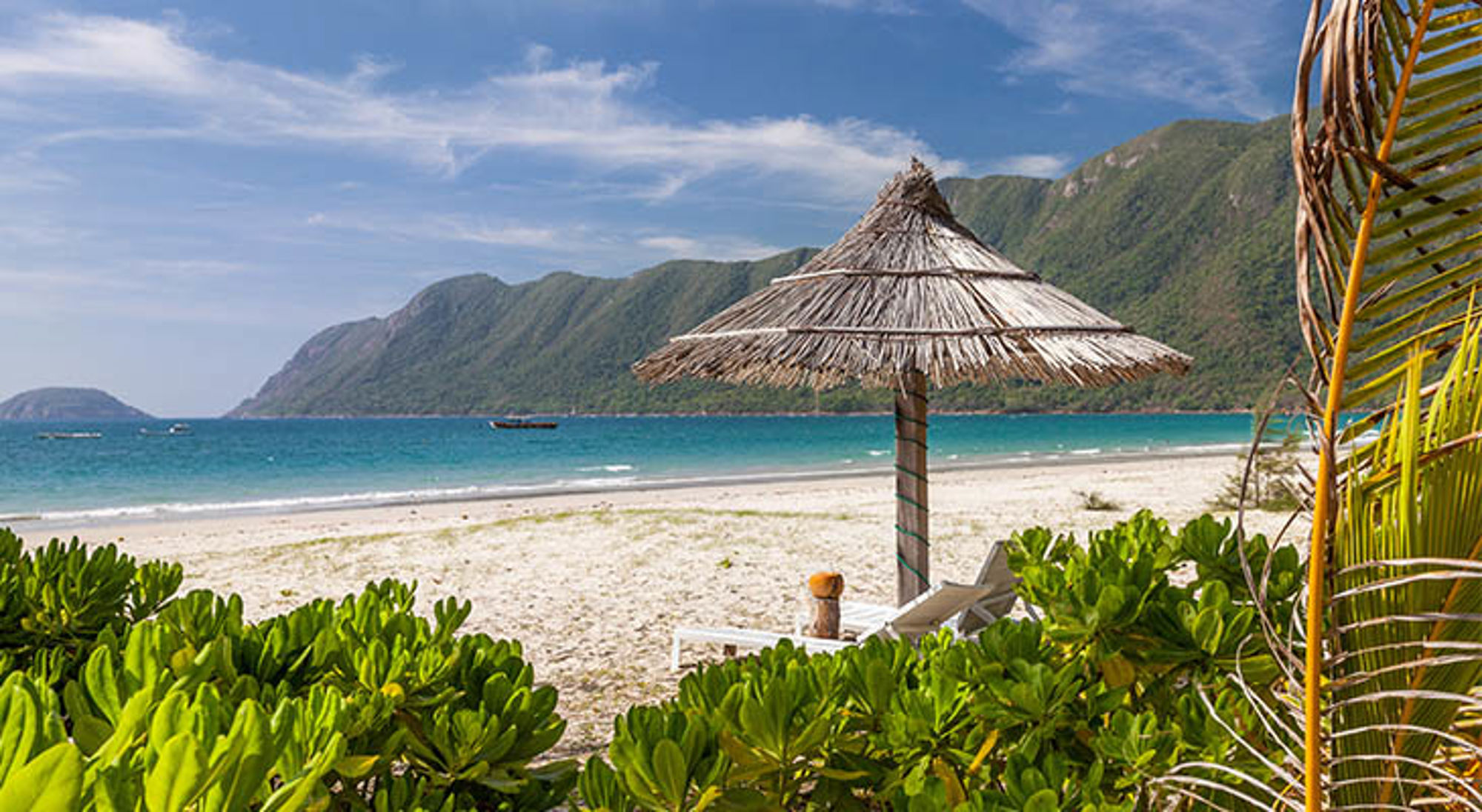 Con Dao Rieten parasol en ligstoel op zandstrand met turquoise zee en groene bergen op de achtergrond in Vietnam.