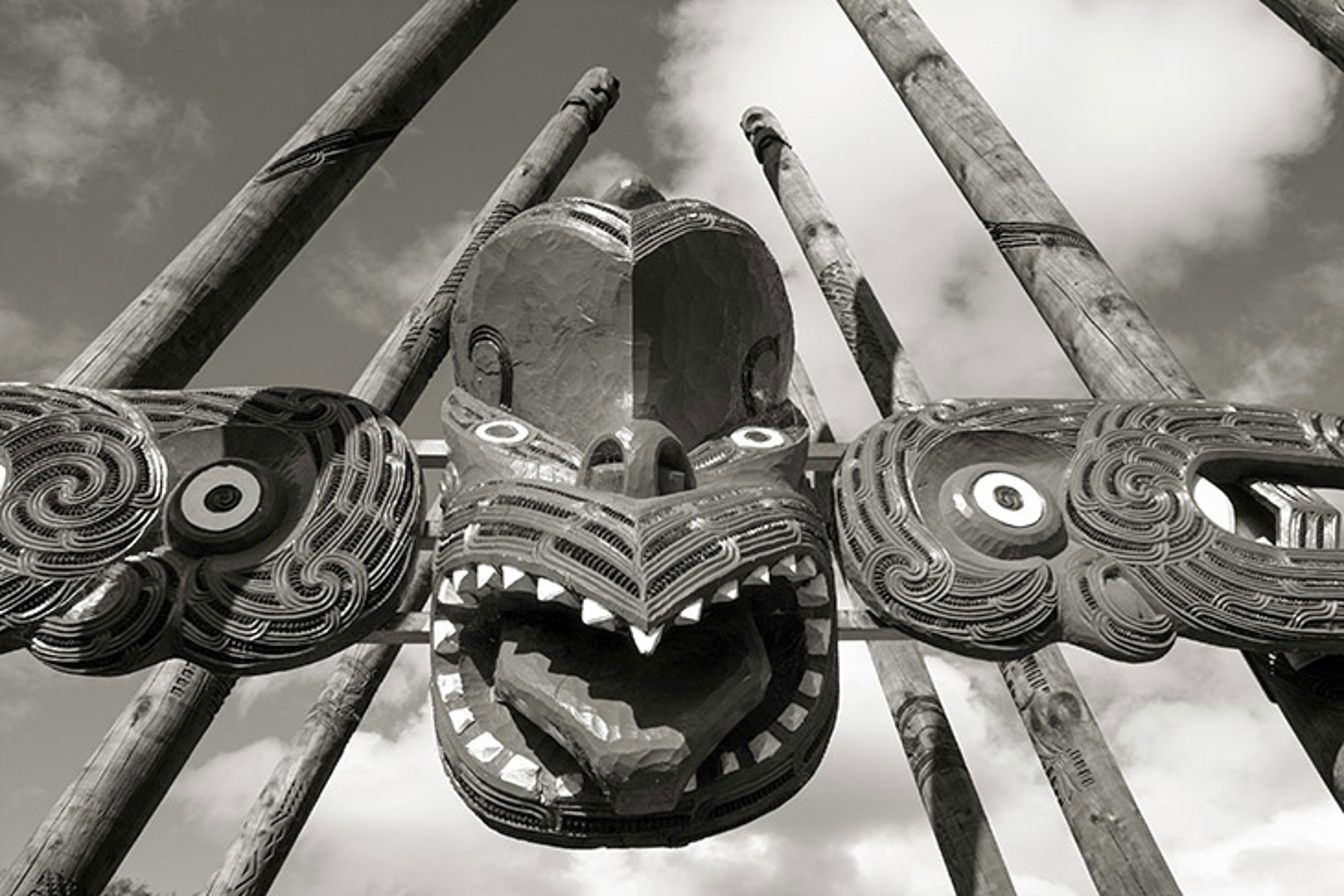 Maori beeld cultuur - Nieuw-Zeeland Houten totemmasker met uitgesneden patronen en tanden, tussen hoge palen tegen een bewolkte lucht.