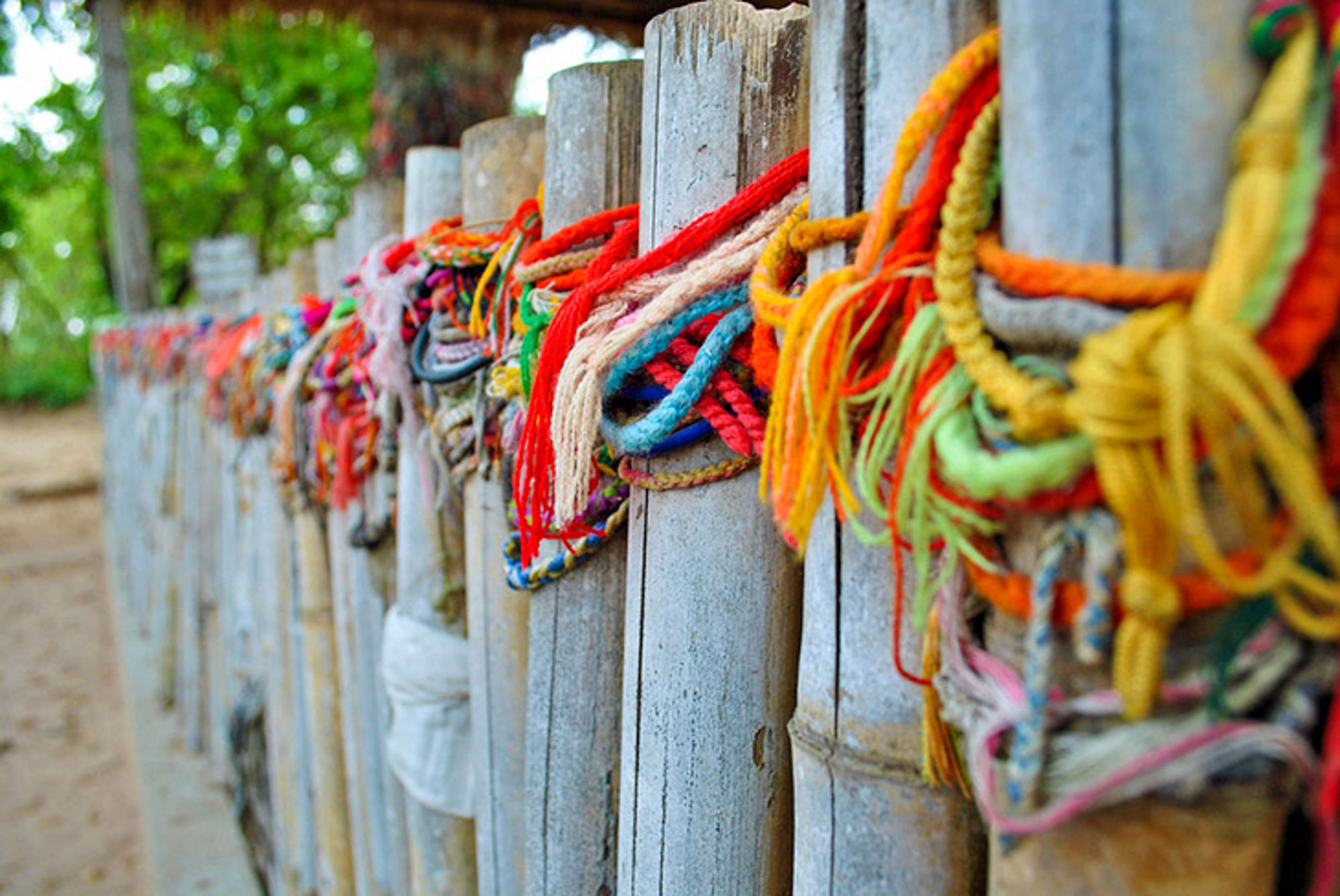Cambodja Kleurrijke touwtjes en linten geknoopt aan een houten hek langs een zandpad.