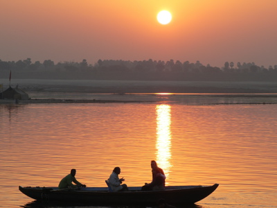 Boot met drie mensen op de Ganges bij zonsopkomst, met gouden weerspiegeling op het water.