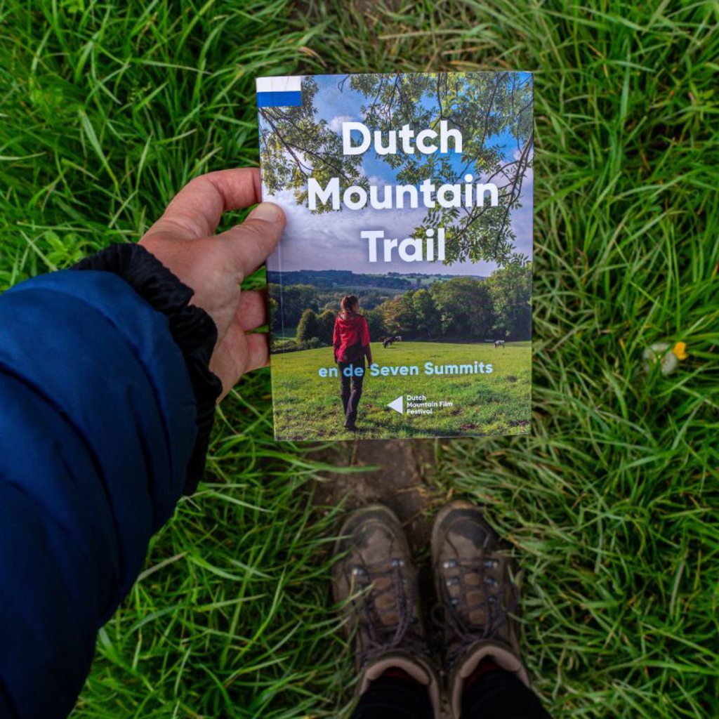Wandelgids Dutch Mountain Trail in hand boven gras, met wandelschoenen in beeld.