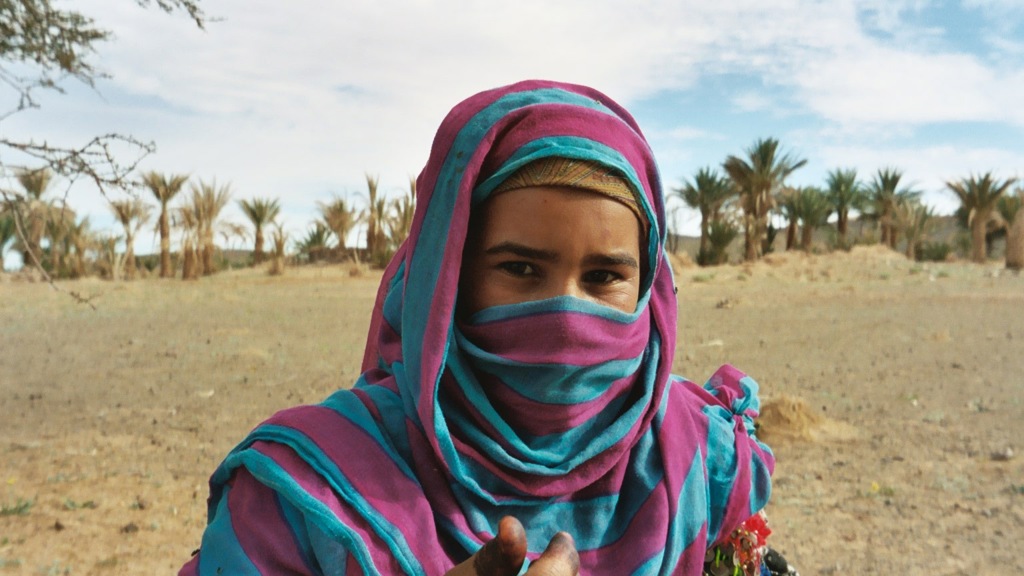 Portret van een vrouw met kleurrijke sjaal in een woestijnlandschap met palmbomen in Marokko.