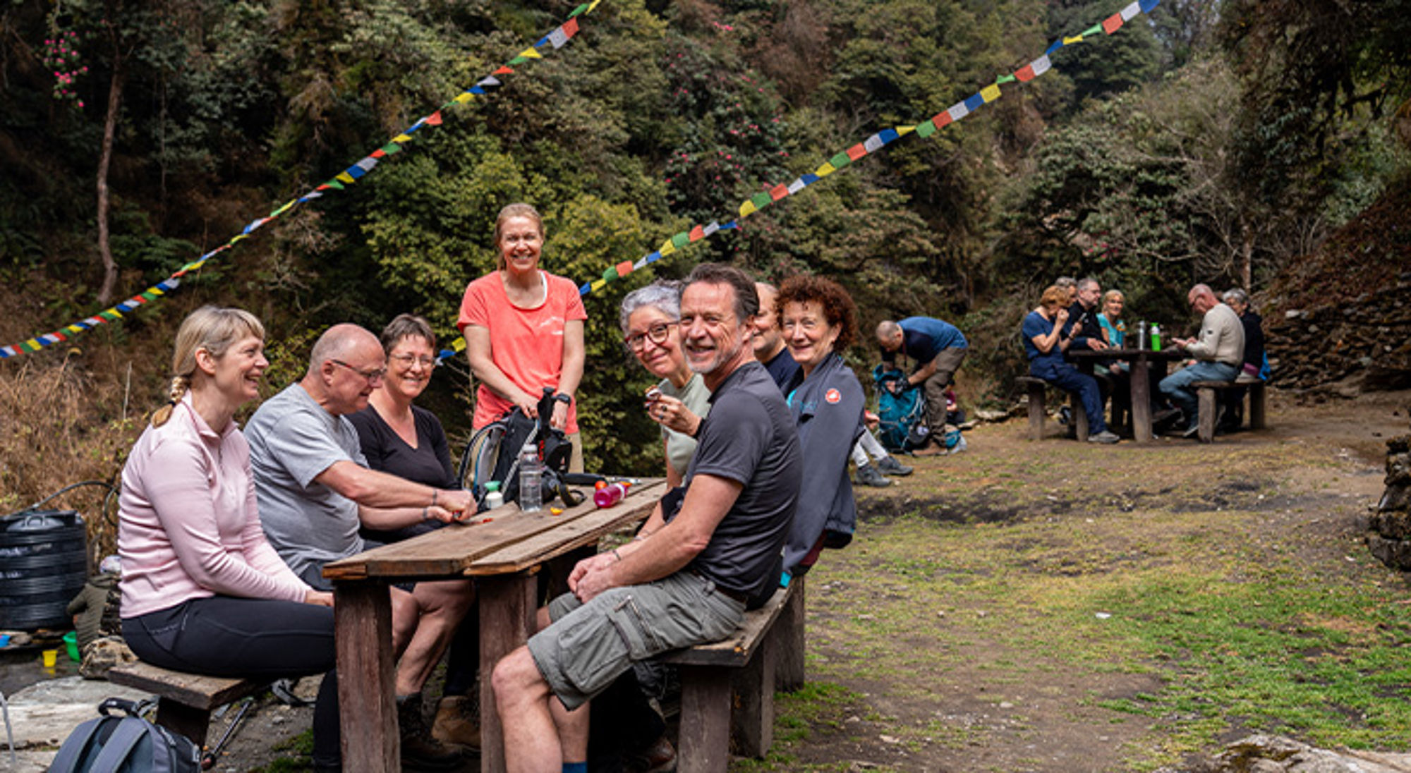 Ghorepani trekking onderweg Wandelaars pauzeren aan picknicktafels in het bos, met kleurrijke vlaggetjes boven het rustpunt.