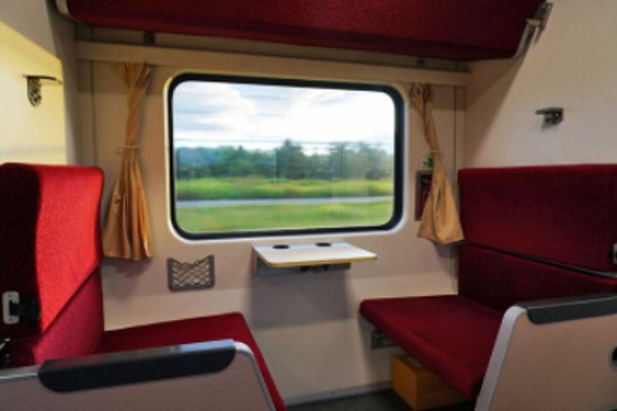 Interieur van een treincompartiment met rode stoelen en uitzicht op groen landschap door het raam.