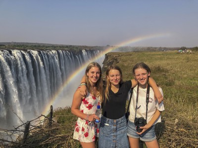 Drie reizigers poseren bij de Victoria Falls met een regenboog in de nevel.