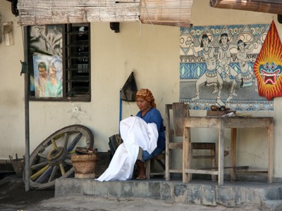 Balinese vrouw zit voor een huis en vouwt witte stof, met traditionele muurschildering op de achtergrond.