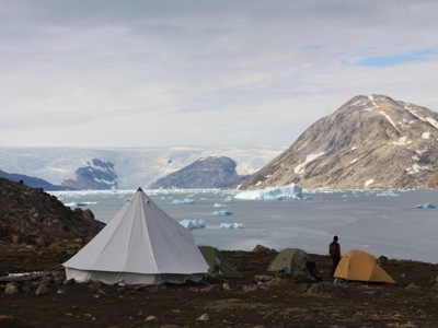 Kampeerplek aan een fjord in Groenland met tenten en drijvende ijsbergen, omringd door bergen.