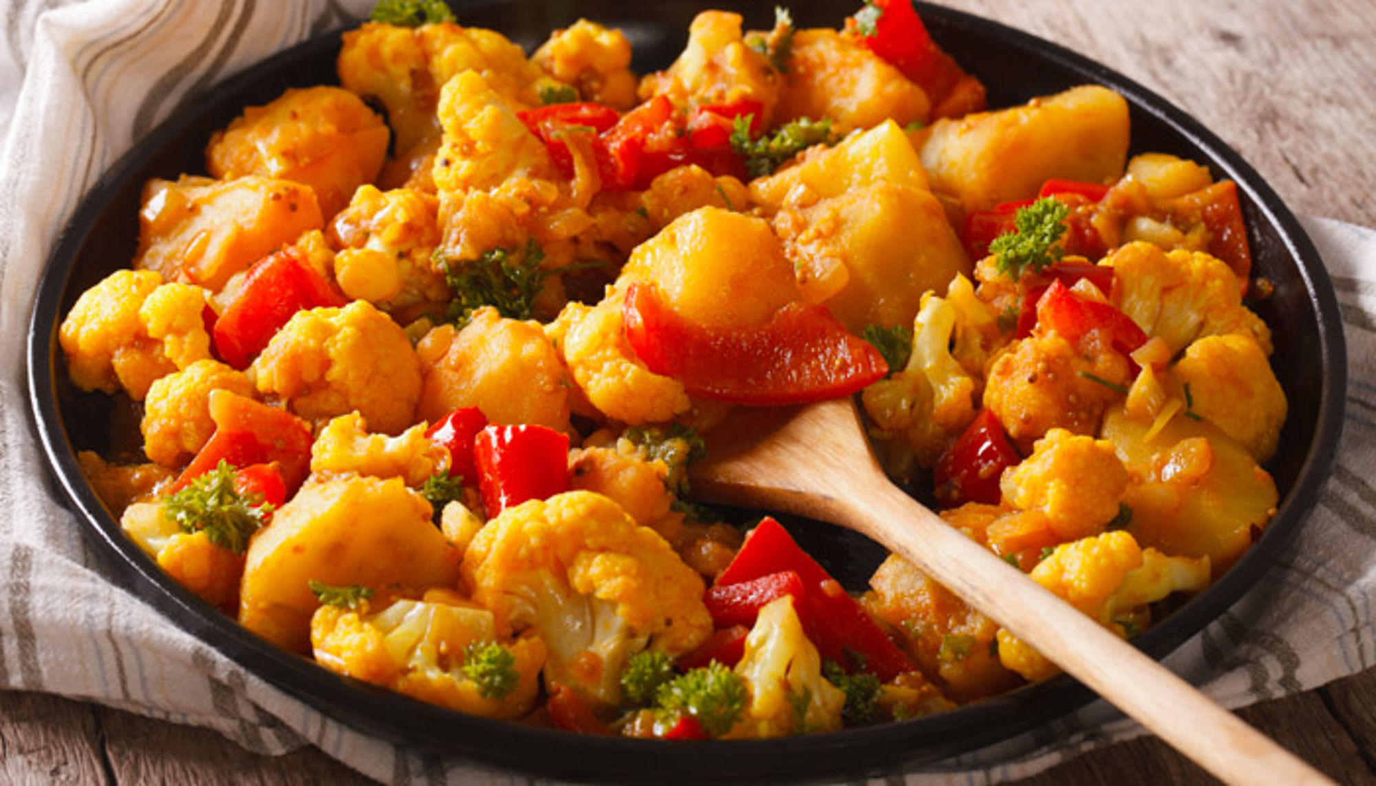 aloo gobi gerecht in india Bloemkoolcurry met aardappel en rode paprika in een pan, geserveerd met een houten lepel.