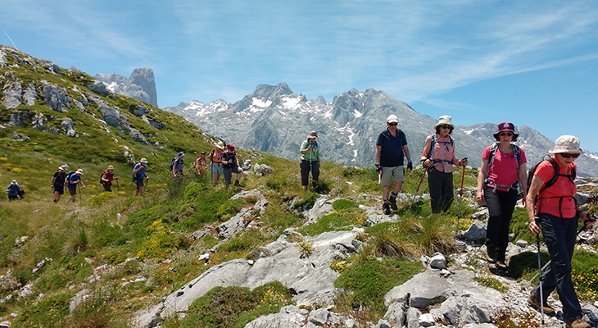 Wandelgroep op bergpad met uitzicht op ruige, besneeuwde bergtoppen onder een blauwe lucht.