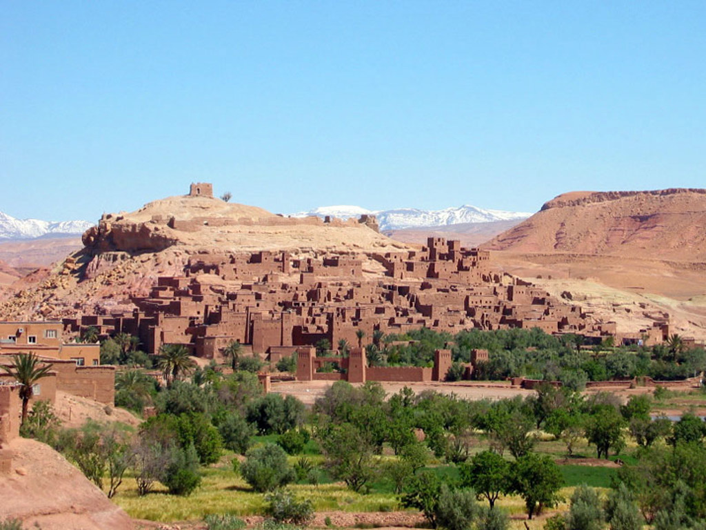 Aït Ben Haddou kasbah in Marokko met lemen huizen, groene oase en besneeuwde Atlasbergen op de achtergrond.