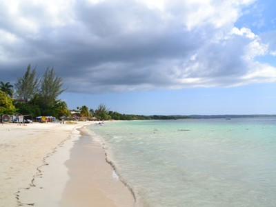 Zandstrand aan de kust van Jamaica met palmbomen, helderblauw zeewater en dreigende wolkenlucht.