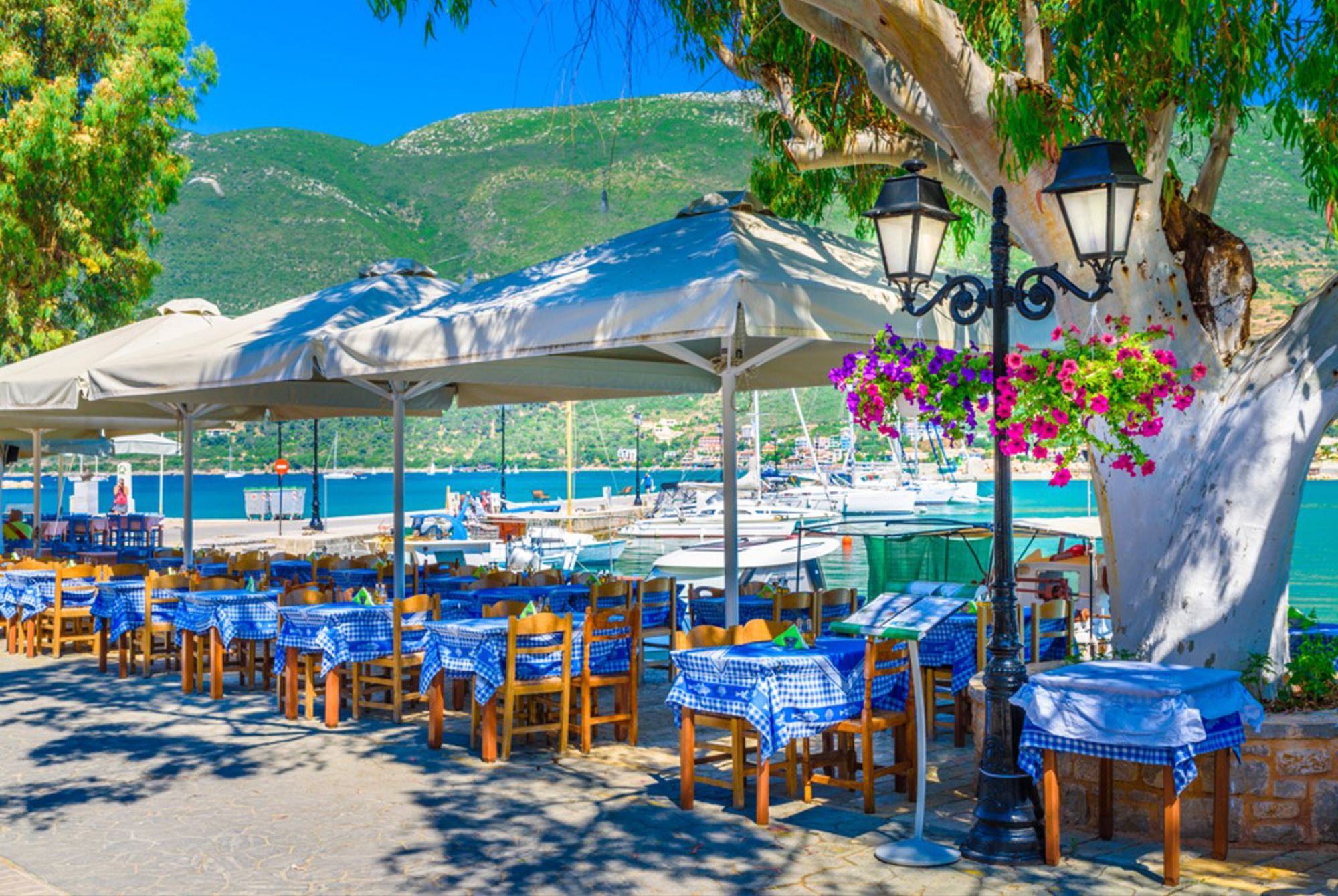Terras aan de haven in Griekenland met tafels onder parasols, jachten op turquoise zee en groene bergen.