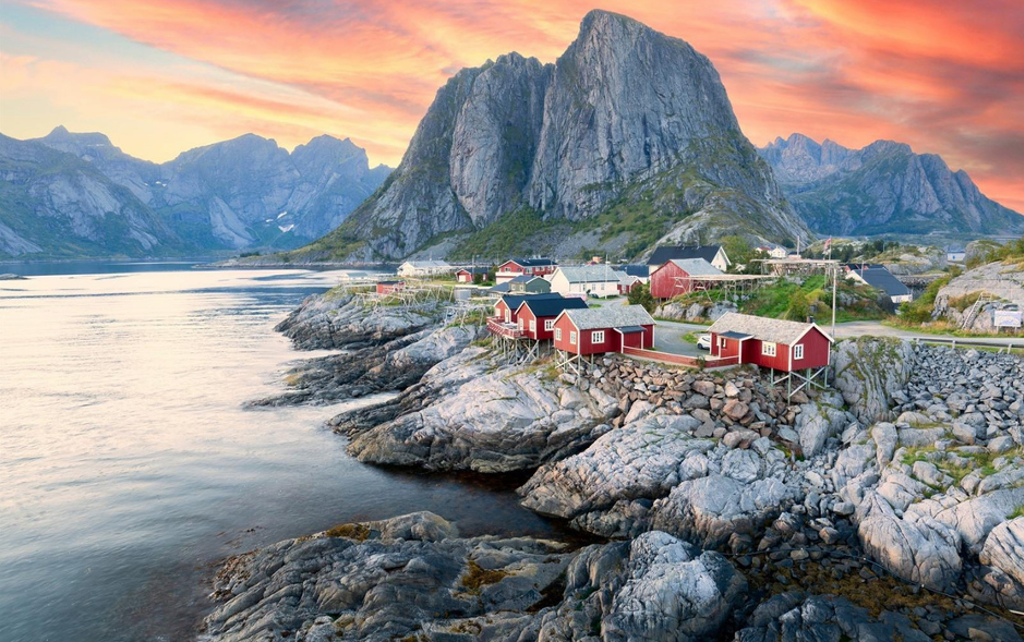 Rode vissershuisjes aan de kust van de Lofoten in Noorwegen, met bergen en zonsondergang op de achtergrond.