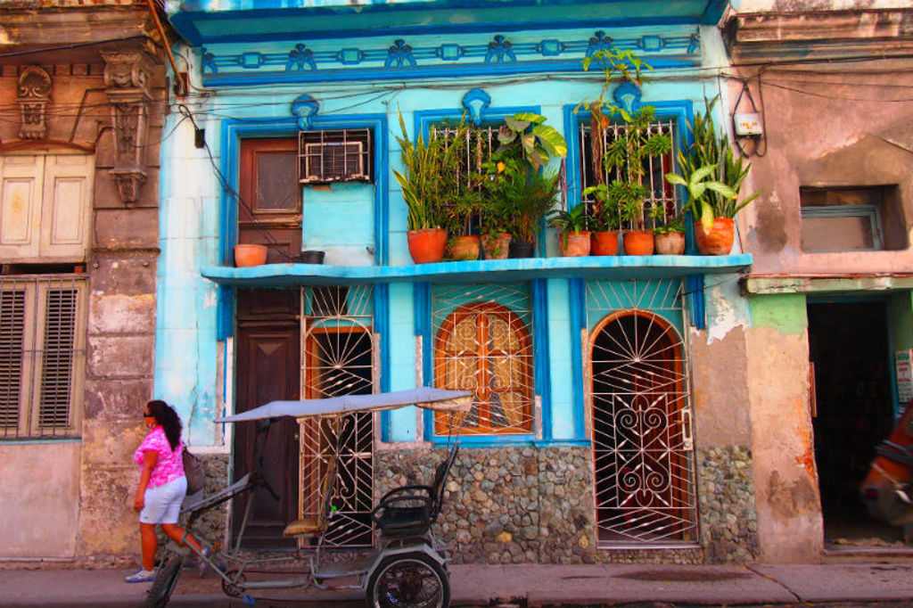 Kleurrijke gevel in Cuba met balkon vol planten en een fietstaxi op straat.
