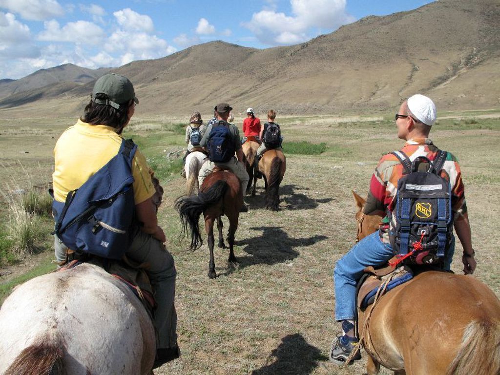 Groepsrondreis Mongolië: reizigers te paard door de uitgestrekte steppe met bergen onder een blauwe lucht.