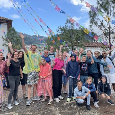 Groepsfoto van gezinnen tijdens familiereis Mexico op een plein met kleurrijke vlaggetjes en bergen op de achtergrond.