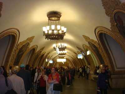 Drukke metrotunnel in Moskou met sierlijke bogen en grote kroonluchters.