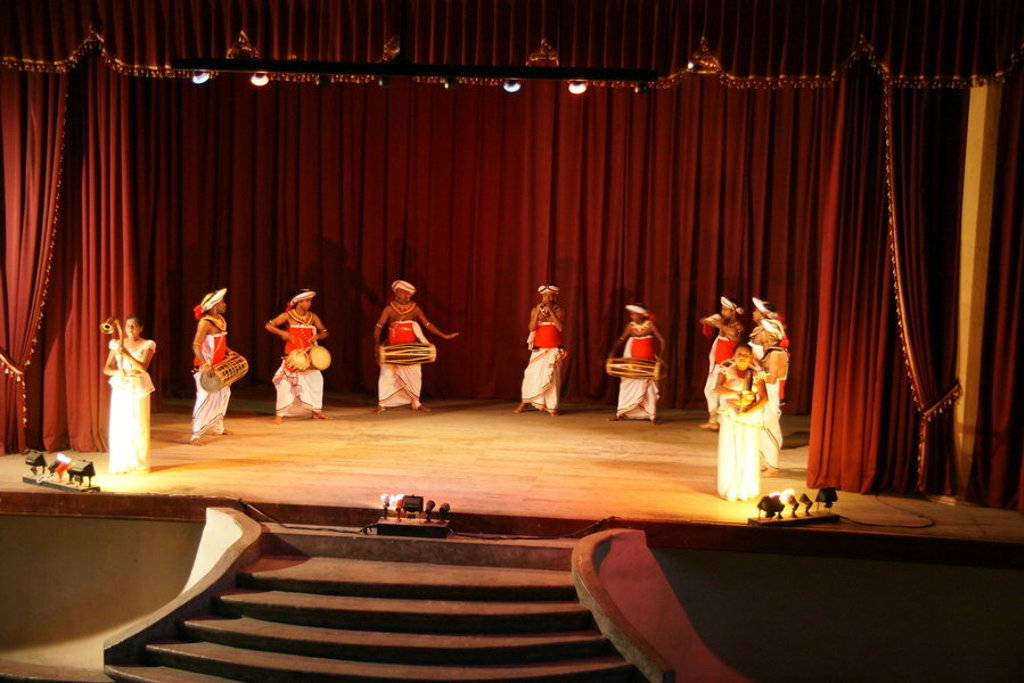 Traditionele Sri Lankaanse dans- en drumvoorstelling op een theaterpodium met rode gordijnen.