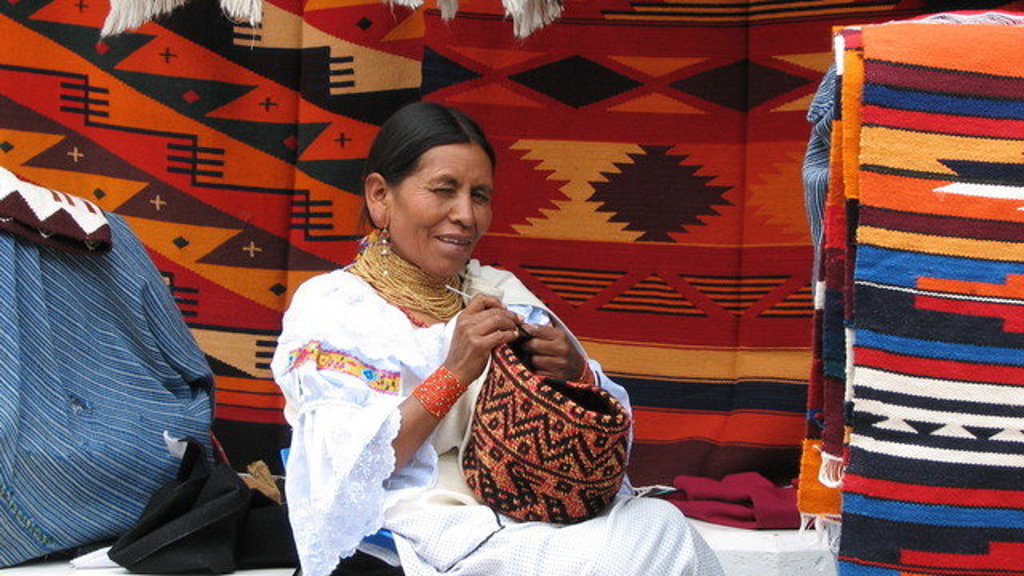 Vrouw in traditionele kleding weeft een tas op een markt in Ecuador, met kleurrijke textiel op de achtergrond.