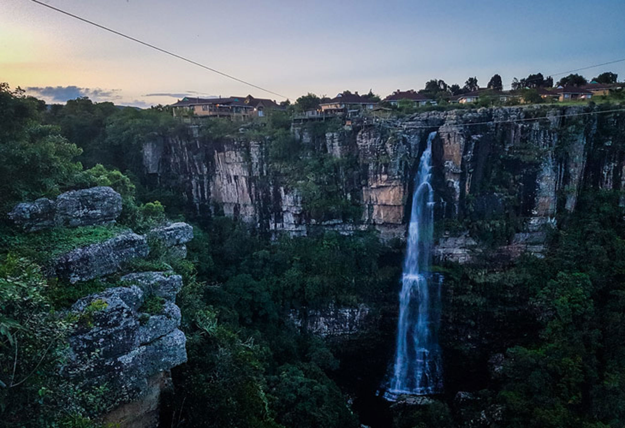 Waterval die van een hoge rotswand in een groene kloof stort, met huizen op de rand bij zonsondergang.