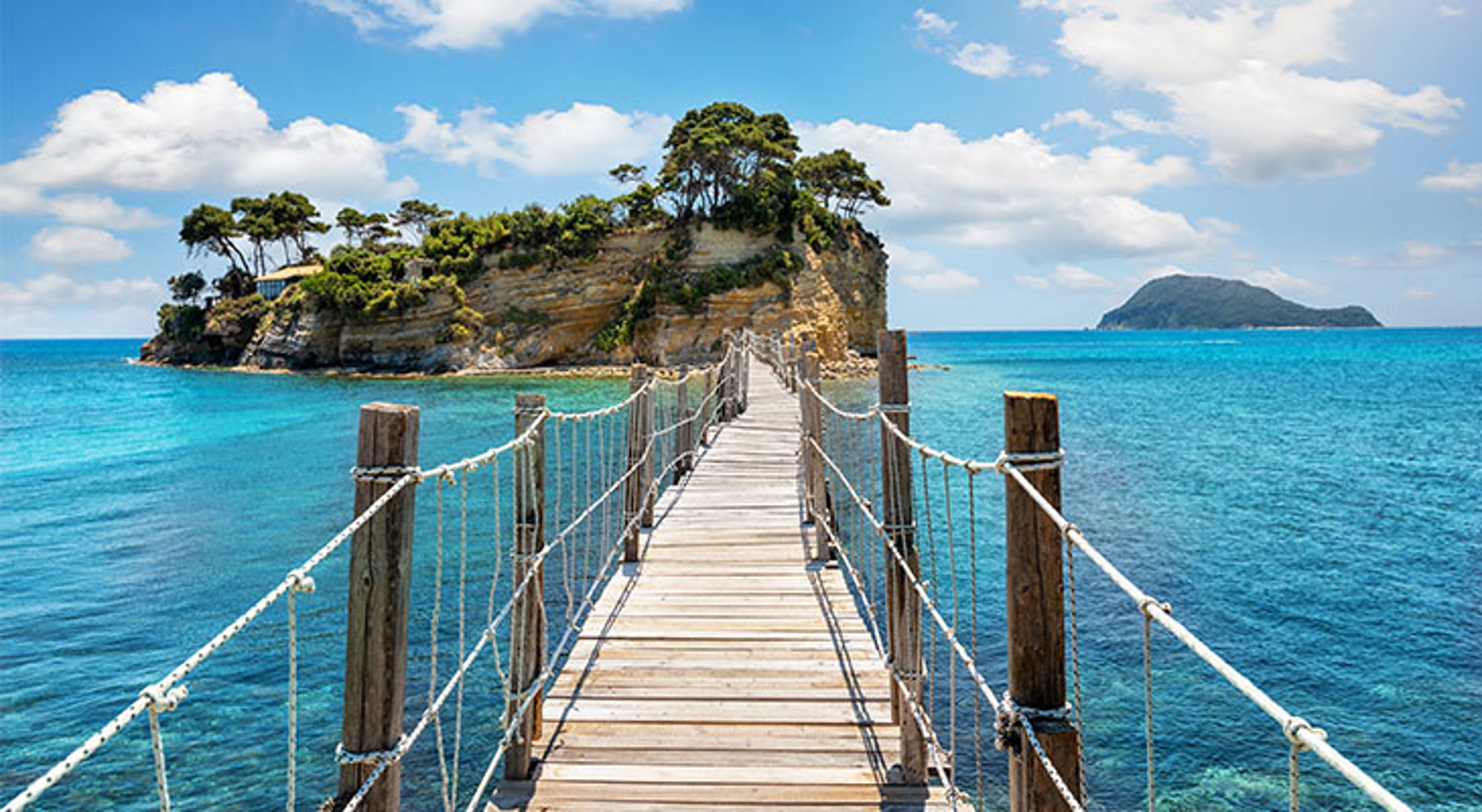 Zakynthos Houten loopbrug over helderblauw water naar een groen eiland onder een blauwe lucht met wolken.